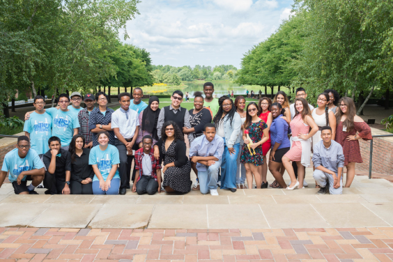 Chicago Botanic Garden Science Career Continuum students pose for a group photo on steps at the Chicago Botanic Garden. Bricks and cement steps in the foreground. Students in the midground. Trees on either side of the background, and a lawn and lake in the background under a cloudy blue sky.
