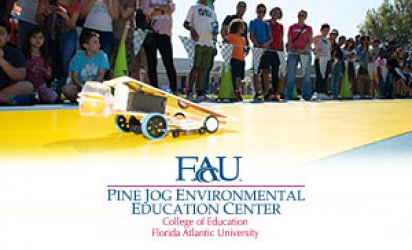 A line of onlookers watch and film as a small solar call shaped to a point races across a yellow track laid out over a blue turf, with triangular flags behind. The Pine Jog logo is in the bottom third over a feathered white backdrop.