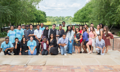 Chicago Botanic Garden Science Career Continuum students pose for a group photo on steps at the Chicago Botanic Garden. Bricks and cement steps in the foreground. Students in the midground. Trees on either side of the background, and a lawn and lake in the background under a cloudy blue sky.