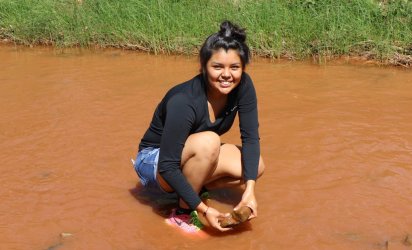 Teenage participant of E4 STEM kneeling in muddy water way with a grassy shore behind her.